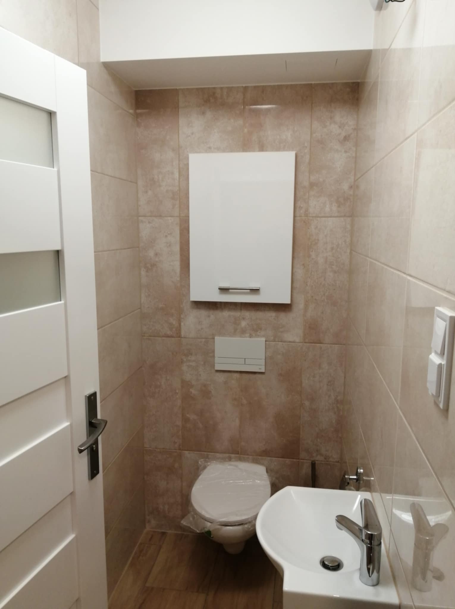 Wykończona łazienka w beżowych płytkach z białą armaturą: umywalka, toaleta z deską pod folią, szafka wisząca. Białe drzwi z klamką po lewej stronie.