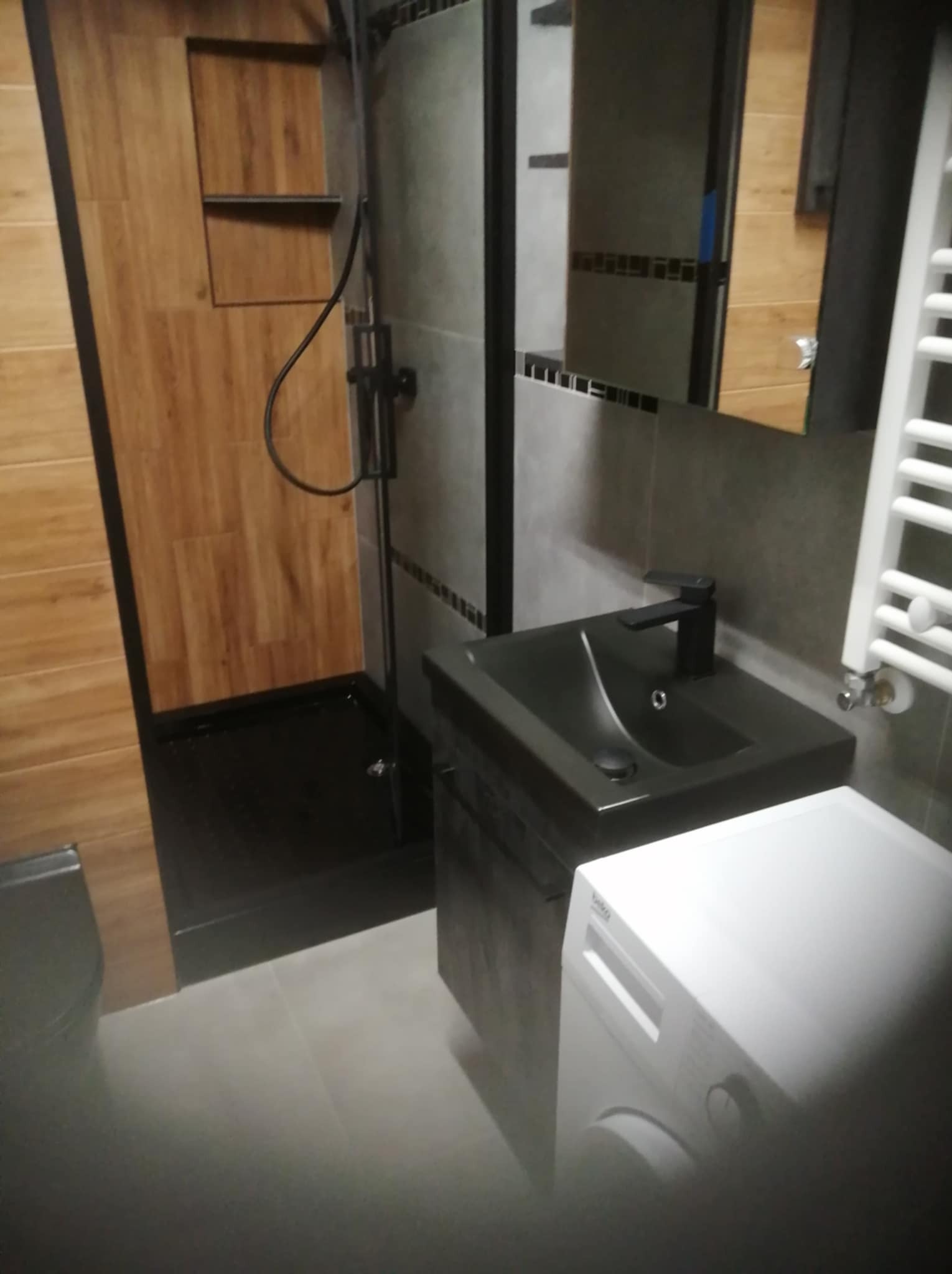 Wykończona łazienka z kabiną prysznicową typu walk-in, umywalką w kolorze czarnym, grzejnikiem drabinkowym i pralką. Drewnopodobne płytki na ścianach kabiny.