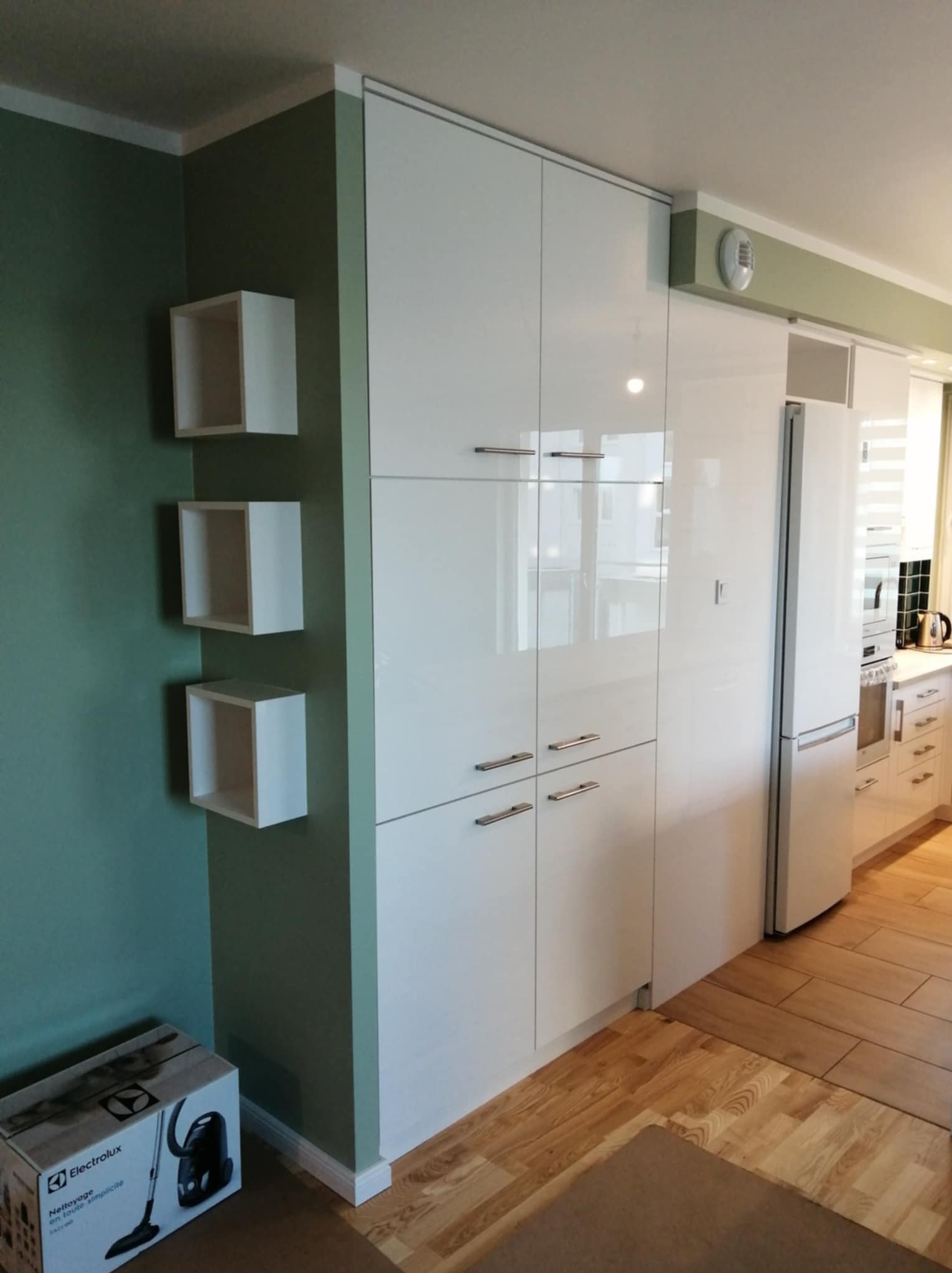 Nowoczesna kuchnia z białymi, lakierowanymi szafkami, trzema białymi półkami kostkami na ścianie i parkietem. W rogu widoczny karton odkurzacza Electrolux.