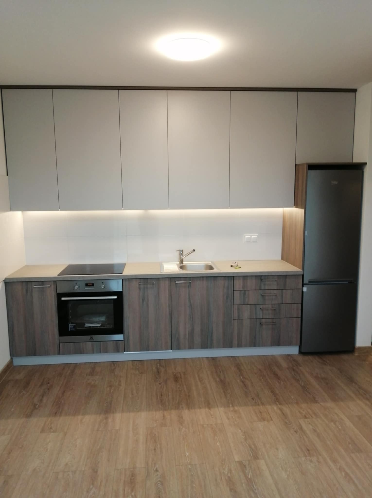 Nowoczesna kuchnia z szarymi szafkami górnymi i dolnymi w odcieniach drewna, oświetleniem LED pod szafkami i czarną lodówką. Jasne panele na podłodze.