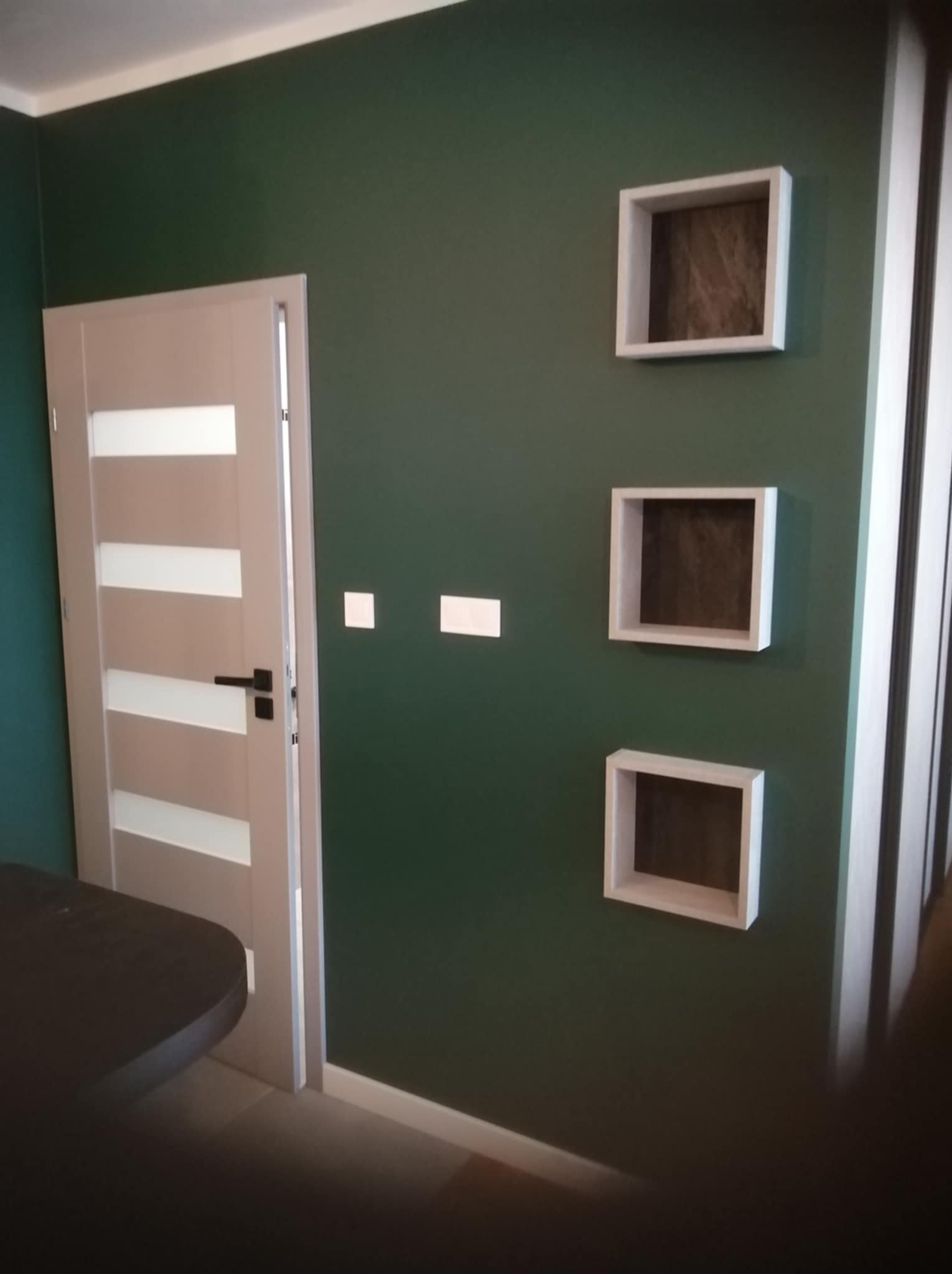 Nowoczesne wnętrze: zielona ściana, białe, minimalistyczne półki, drzwi z przeszkleniami i fragment ciemnego blatu. Wykończenie po remoncie.