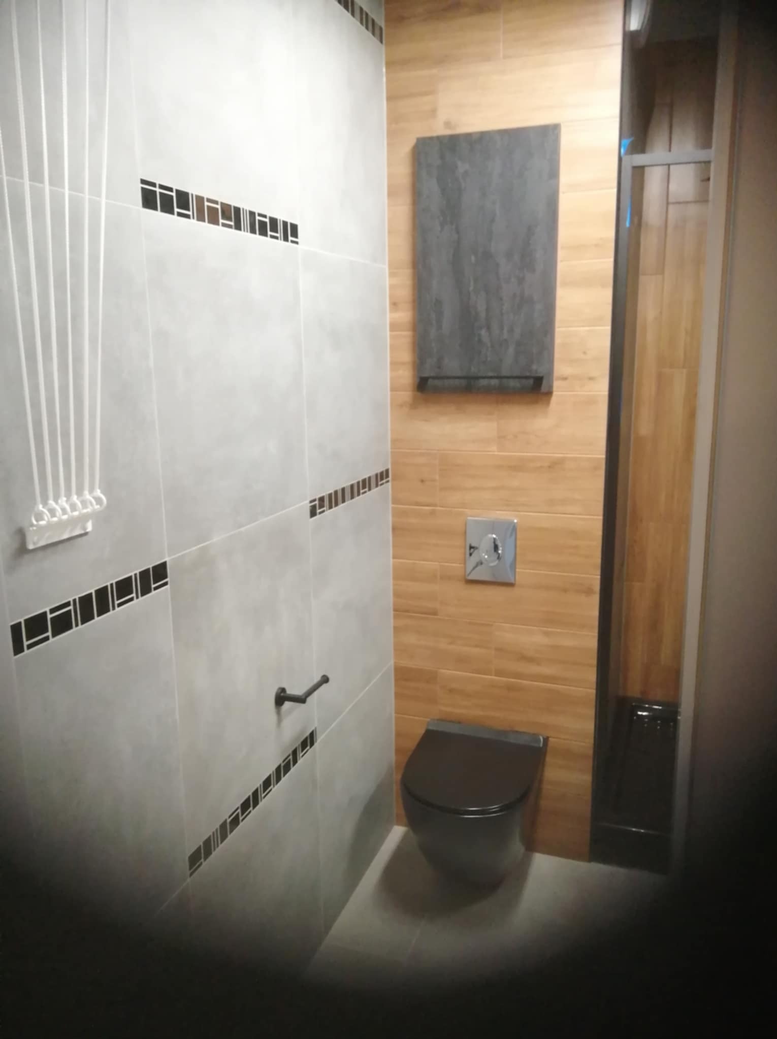 Nowoczesna łazienka w szaro-drewnianej tonacji: szare płytki z dekorami, drewnopodobne płytki, wisząca szafka, czarna toaleta i kabina prysznicowa.