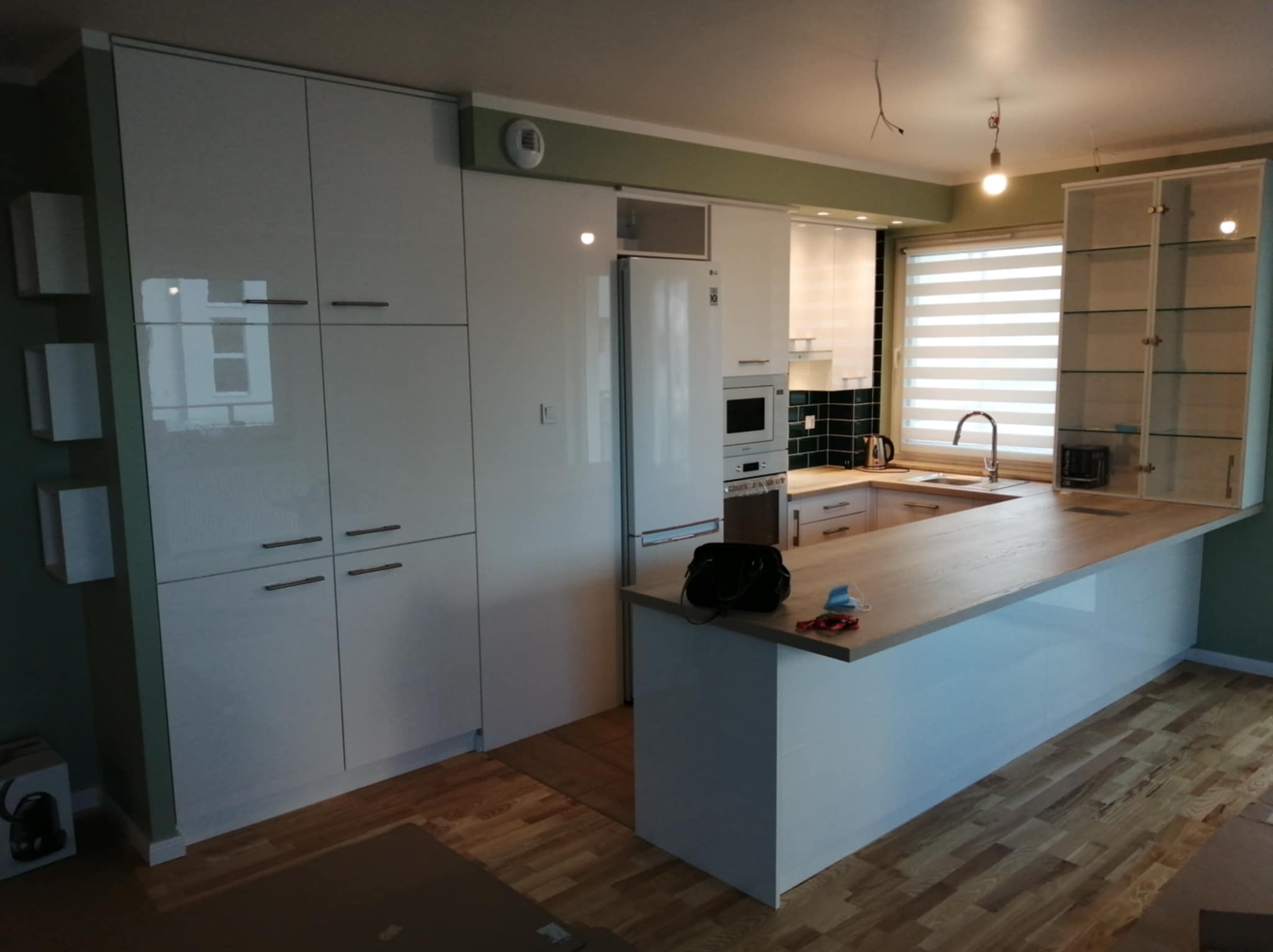 Nowoczesna kuchnia z białymi, lakierowanymi frontami szafek, drewnianym blatem i elementami szklanymi. Widoczny sprzęt AGD i drewniana podłoga.