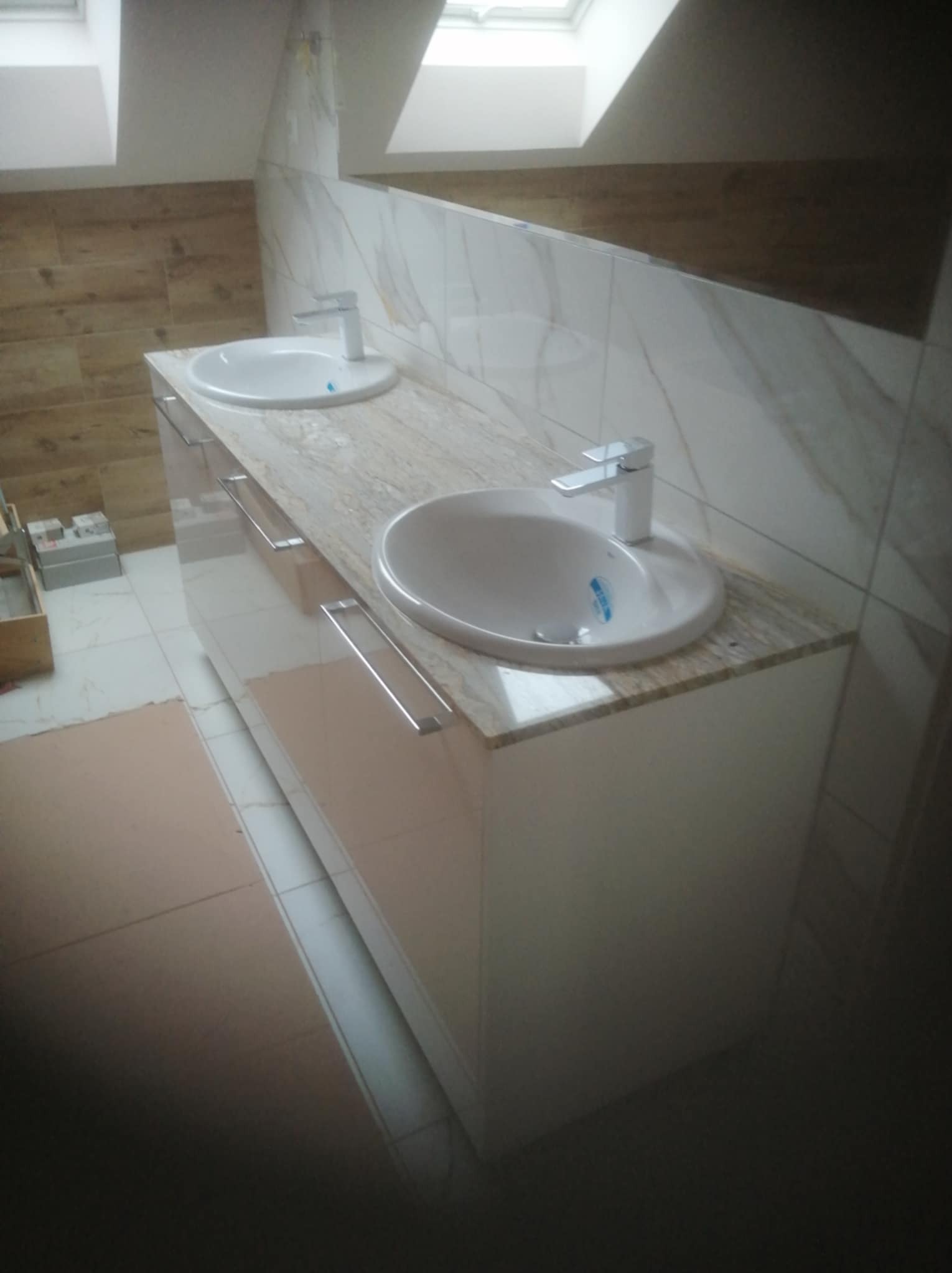 Ukończona łazienka na poddaszu: podwójna umywalka nablatowa z marmurowym blatem i błyszczącymi, kremowymi szafkami. Widoczne okno dachowe i płytki imitujące drewno.