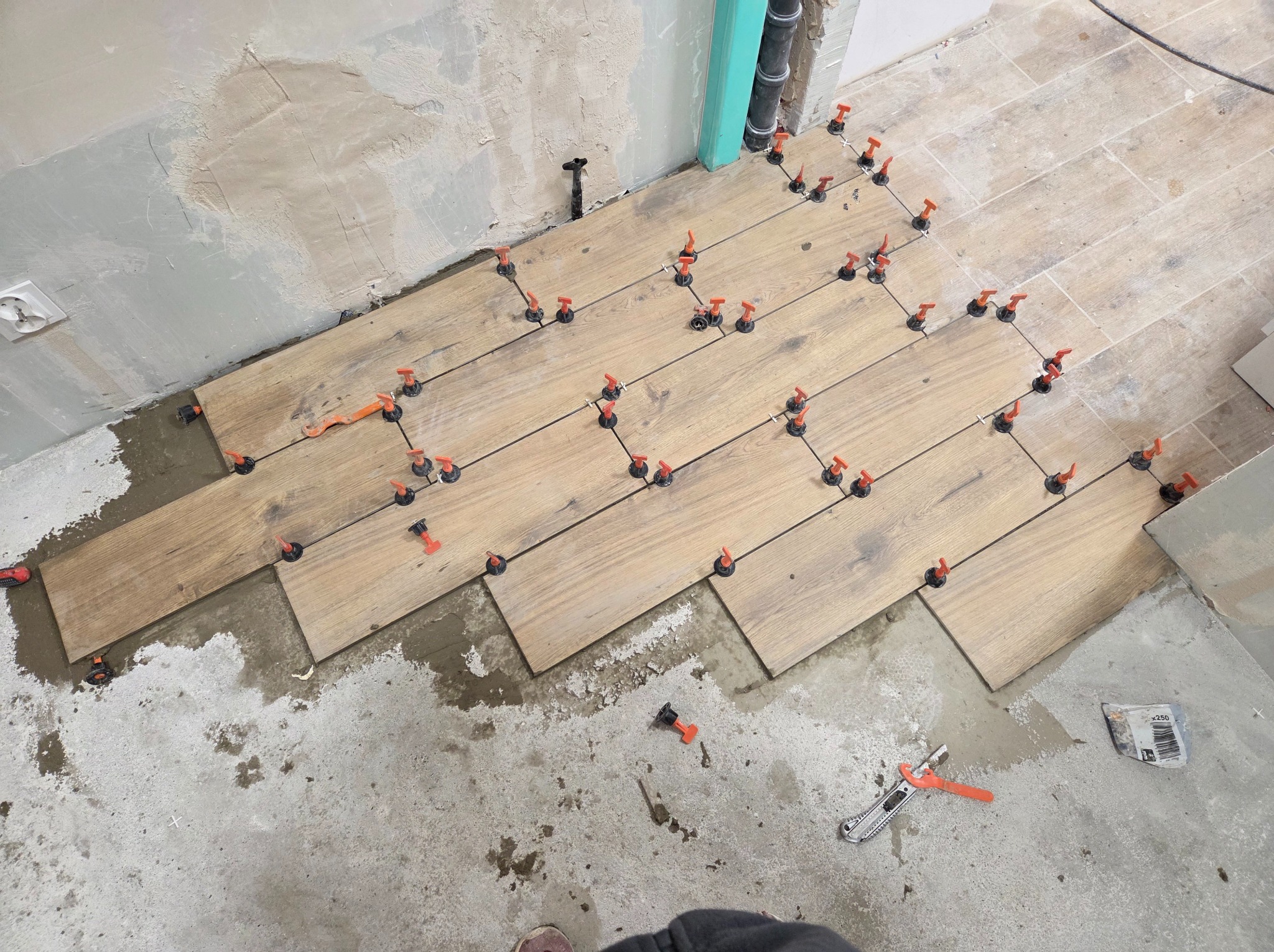 Układanie drewnopodobnych płytek na podłodze z użyciem systemu poziomowania. Widoczne narzędzia glazurnicze i fragment ściany z pozostałościami po szpachlowaniu.