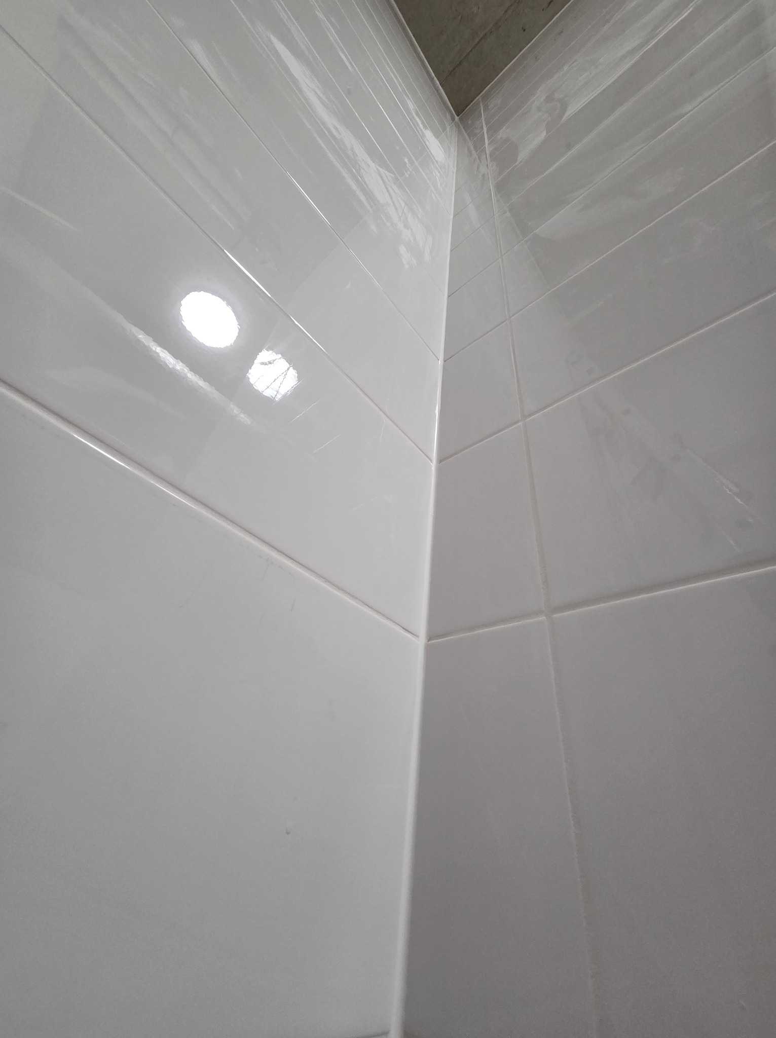 Błyszczące, białe płytki ceramiczne ułożone w rogu łazienki, z widocznym fragmentem surowego sufitu. Minimalistyczny design, jasne wnętrze.