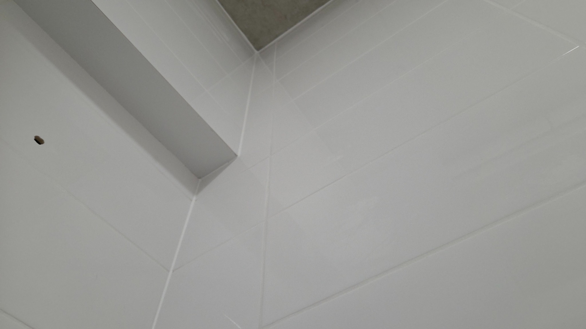 Białe, błyszczące płytki ceramiczne ułożone na ścianie w rogu pomieszczenia, widoczne fugi i fragment sufitu. Minimalistyczne wykończenie wnętrza.