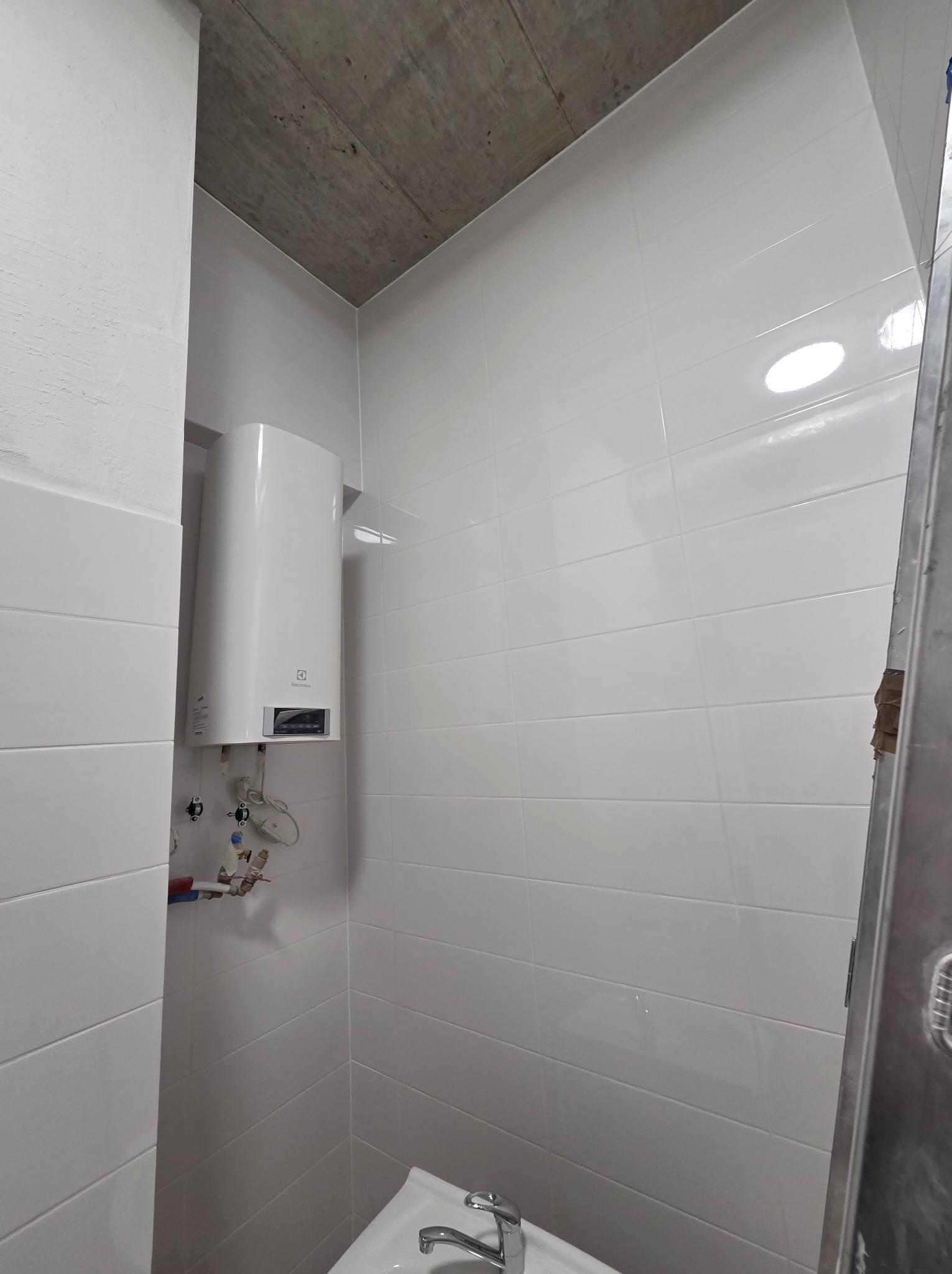 Biała łazienka z płytkami, bojlerem Electrolux i umywalką. Widoczny surowy betonowy sufit i fragment drabiny. Minimalistyczne wykończenie.