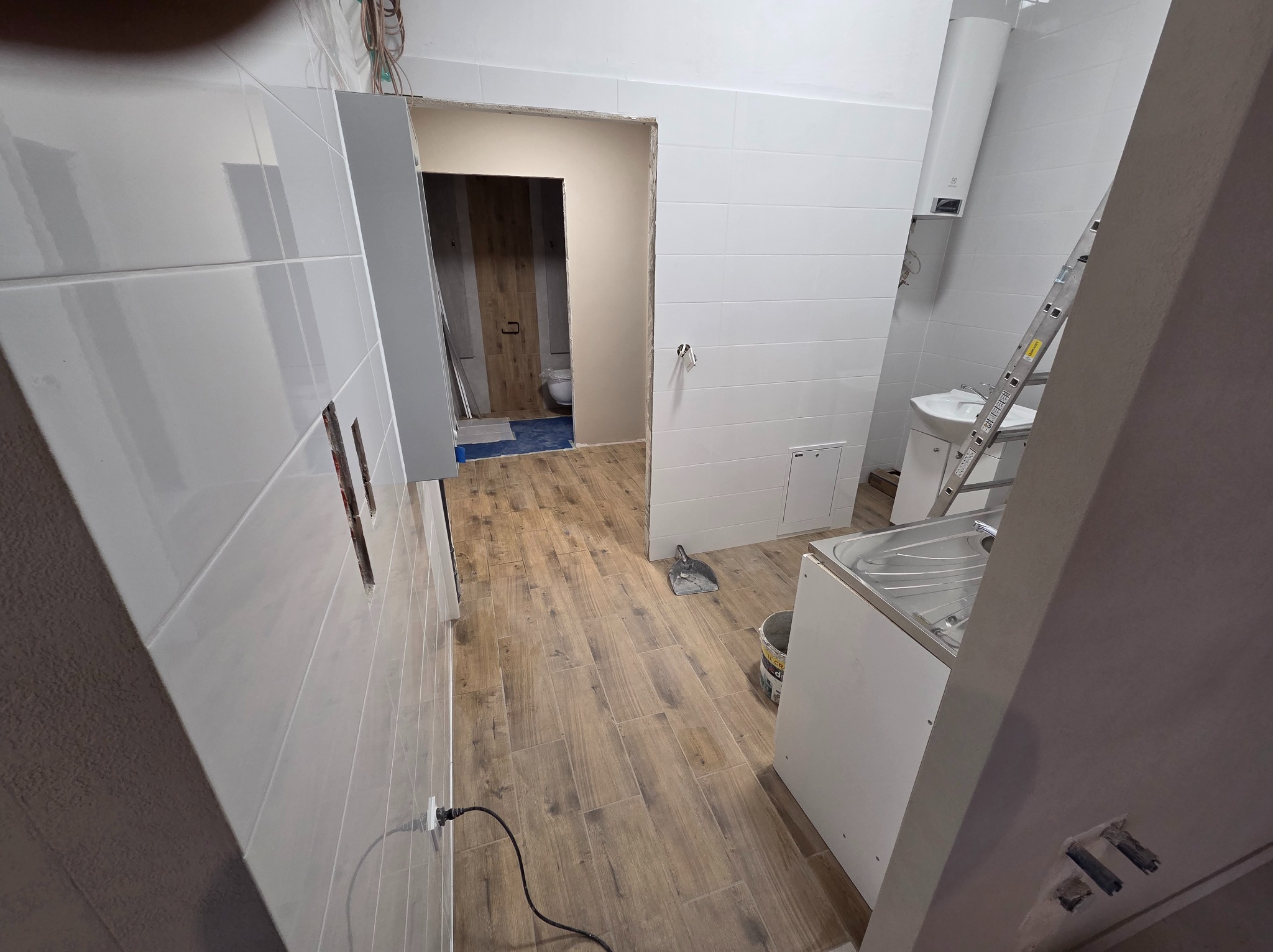 Wnętrze łazienki w trakcie prac wykończeniowych: białe płytki na ścianach, drewnopodobna podłoga, drabina i umywalka. Widoczne niedokończone elementy instalacji.