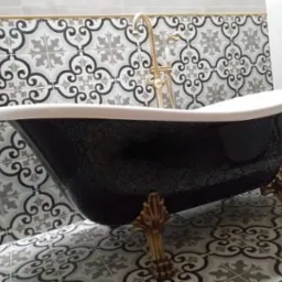 Czarna wanna wolnostojąca na złotych nóżkach, z baterią w stylu retro, na tle ściany i podłogi wyłożonej wzorzystymi, szaro-białymi płytkami.