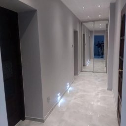 DrigoKomplex B.R.T - Wykończony korytarz w szarościach z podświetlaną listwą przypodłogową i lustrzanymi drzwiami przesuwnymi, widoczne płytki podłogowe i oświetlenie sufitowe.