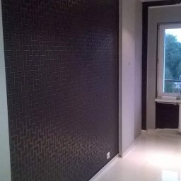 DrigoKomplex B.R.T - Ciemna ściana z geometrycznym wzorem w pokoju z jasną podłogą i oknem. Widoczne gniazdka elektryczne i fragment grzejnika.