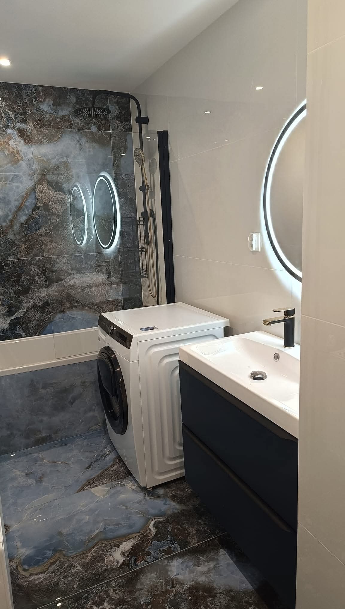 Nowoczesna łazienka z marmurowymi płytkami, okrągłym lustrem z podświetleniem LED, pralką i umywalką z szafką w kolorze granatowym. Bateria w kolorze czarnym.