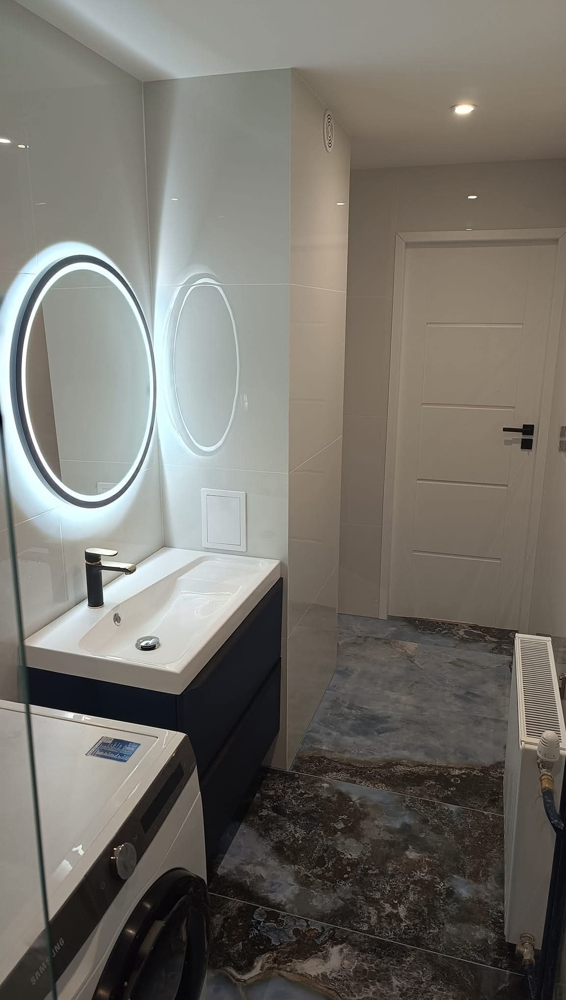 Nowoczesna łazienka z umywalką, okrągłym lustrem z podświetleniem LED, pralką i płytkami imitującymi marmur na podłodze.