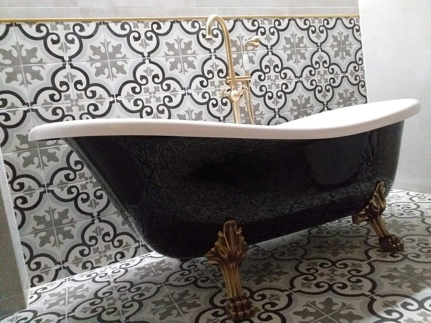 Czarna wanna wolnostojąca na złotych nóżkach, z baterią w stylu retro, na tle ściany i podłogi wyłożonej wzorzystymi, szaro-białymi płytkami.