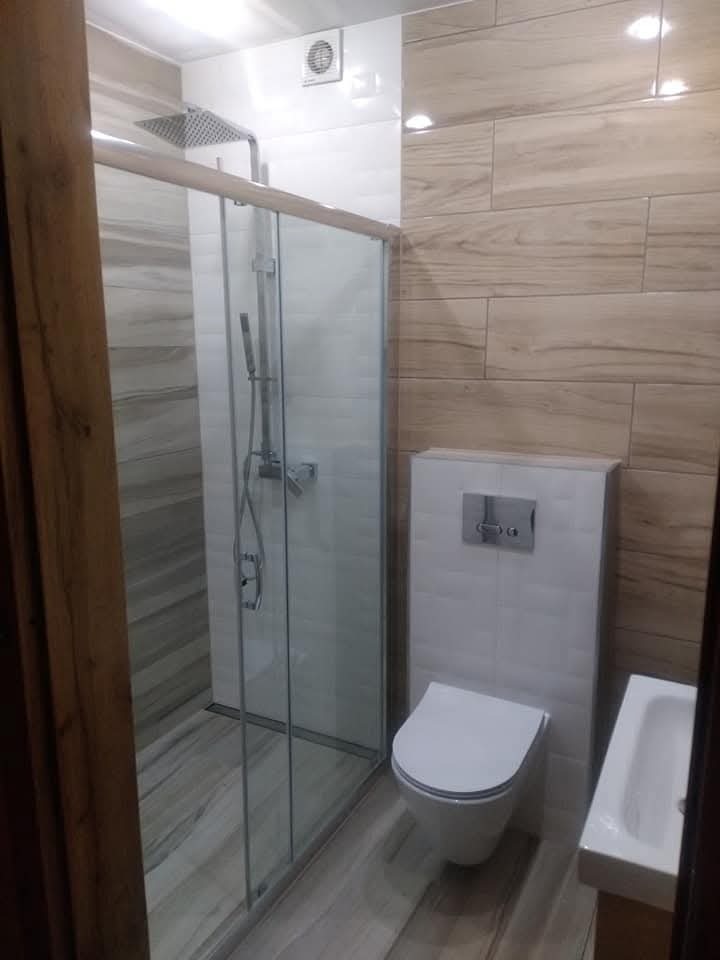 Nowoczesna łazienka po remoncie: kabina walk-in z deszczownicą, toaleta podwieszana i umywalka nablatowa. Płytki imitujące drewno na ścianie i podłodze.