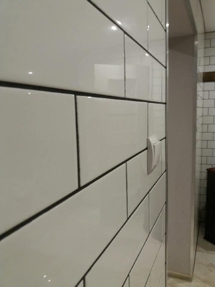 Białe, błyszczące płytki ułożone w cegiełkę z ciemną fugą, widoczne z boku, z włącznikiem światła i fragmentem wnętrza w tle.