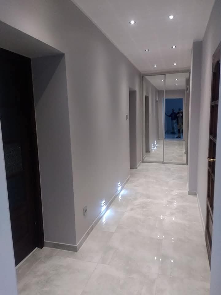 Wykończony korytarz w szarościach z podświetlaną listwą przypodłogową i lustrzanymi drzwiami przesuwnymi, widoczne płytki podłogowe i oświetlenie sufitowe.