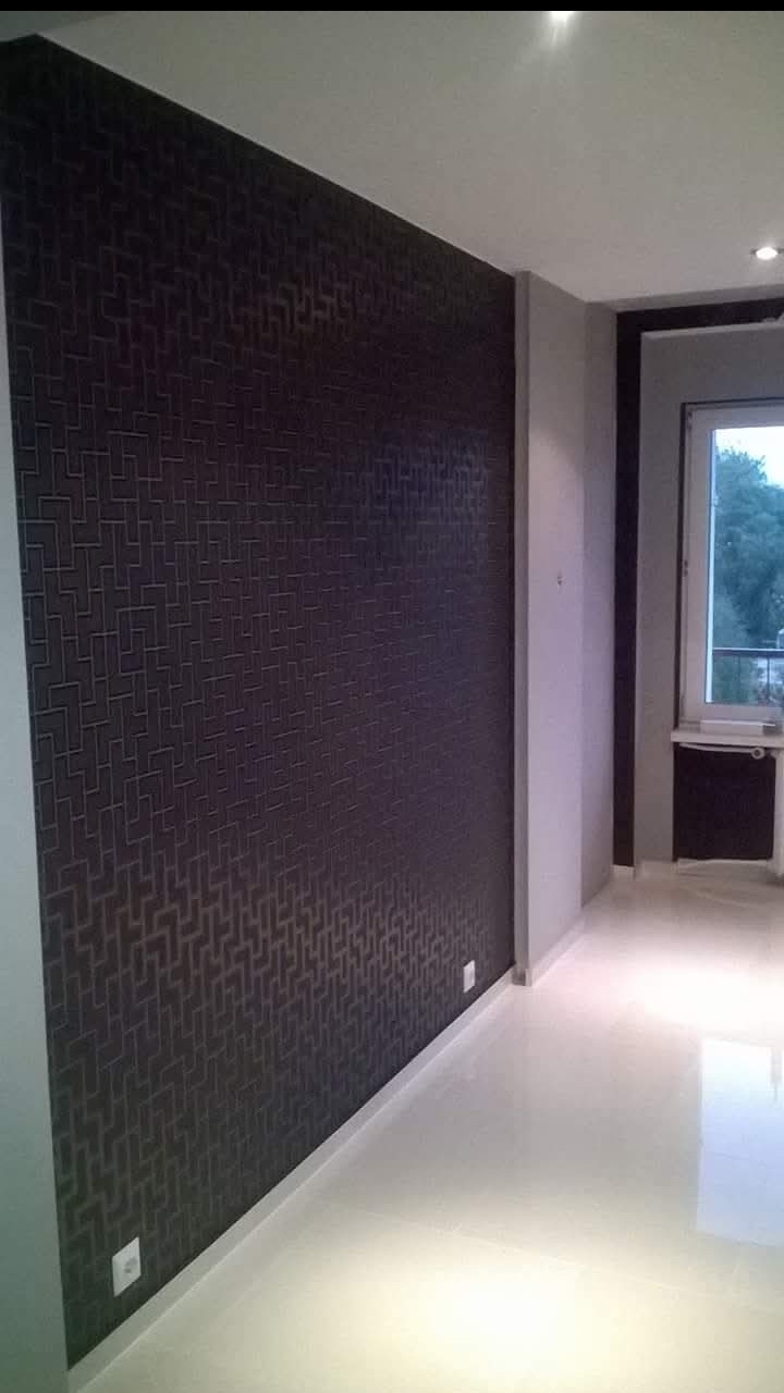 Ciemna ściana z geometrycznym wzorem w pokoju z jasną podłogą i oknem. Widoczne gniazdka elektryczne i fragment grzejnika.