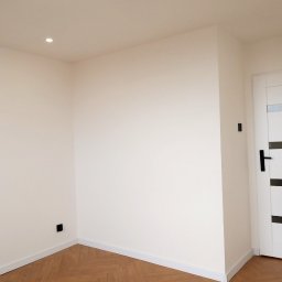 Andrzej Twaróg - Jasne wnętrze po remoncie: świeżo pomalowane ściany w odcieniach bieli, parkiet ułożony w jodełkę i białe drzwi z czarnymi akcentami. Minimalistyczny design.