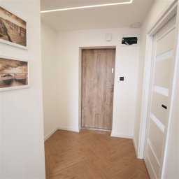 Andrzej Twaróg - Jasne wnętrze przedpokoju z drewnianą podłogą w jodełkę, dwoje drzwi (zewnętrzne i wewnętrzne) oraz dekoracje ścienne. Oświetlenie LED na suficie.