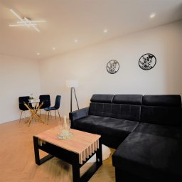 Andrzej Twaróg - Nowoczesny salon z czarną sofą narożną, szklanym stolikiem i dekoracjami ściennymi. Parkiet w jodełkę, lampa podłogowa i sufitowe oświetlenie punktowe.