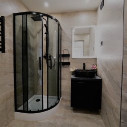 Andrzej Twaróg - Nowoczesna łazienka z kabiną prysznicową w czarnej ramie, umywalką na szafce i podświetlanym lustrem. Beżowe płytki imitujące marmur na ścianach i podłodze.