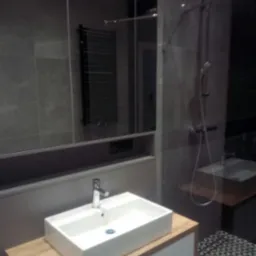 Nowoczesna łazienka: biała, prostokątna umywalka nablatowa, szare płytki, geometryczny wzór na podłodze, lustro z wbudowanym oświetleniem i grzejnik drabinkowy.