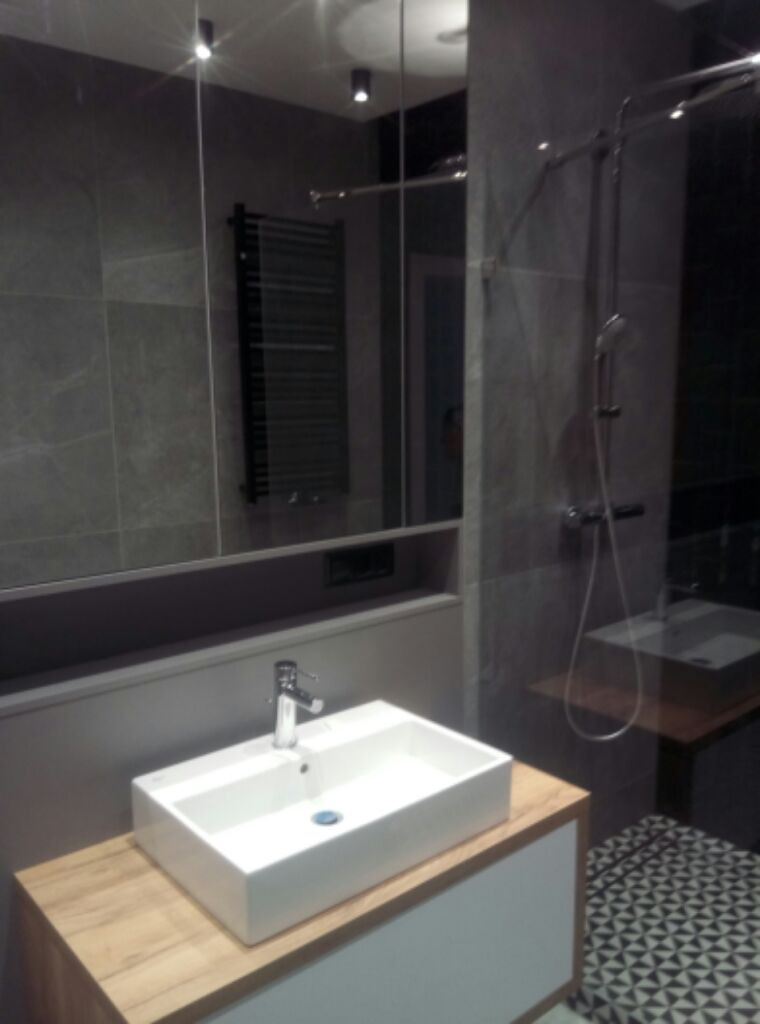 Nowoczesna łazienka: biała, prostokątna umywalka nablatowa, szare płytki, geometryczny wzór na podłodze, lustro z wbudowanym oświetleniem i grzejnik drabinkowy.