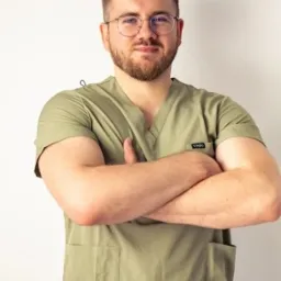 Młody mężczyzna w okularach i oliwkowej bluzie medycznej, stoi na tle jasnej ściany, z założonymi rękami. Uśmiechnięty, z krótką brodą i starannie przystrzyżonymi włosami.