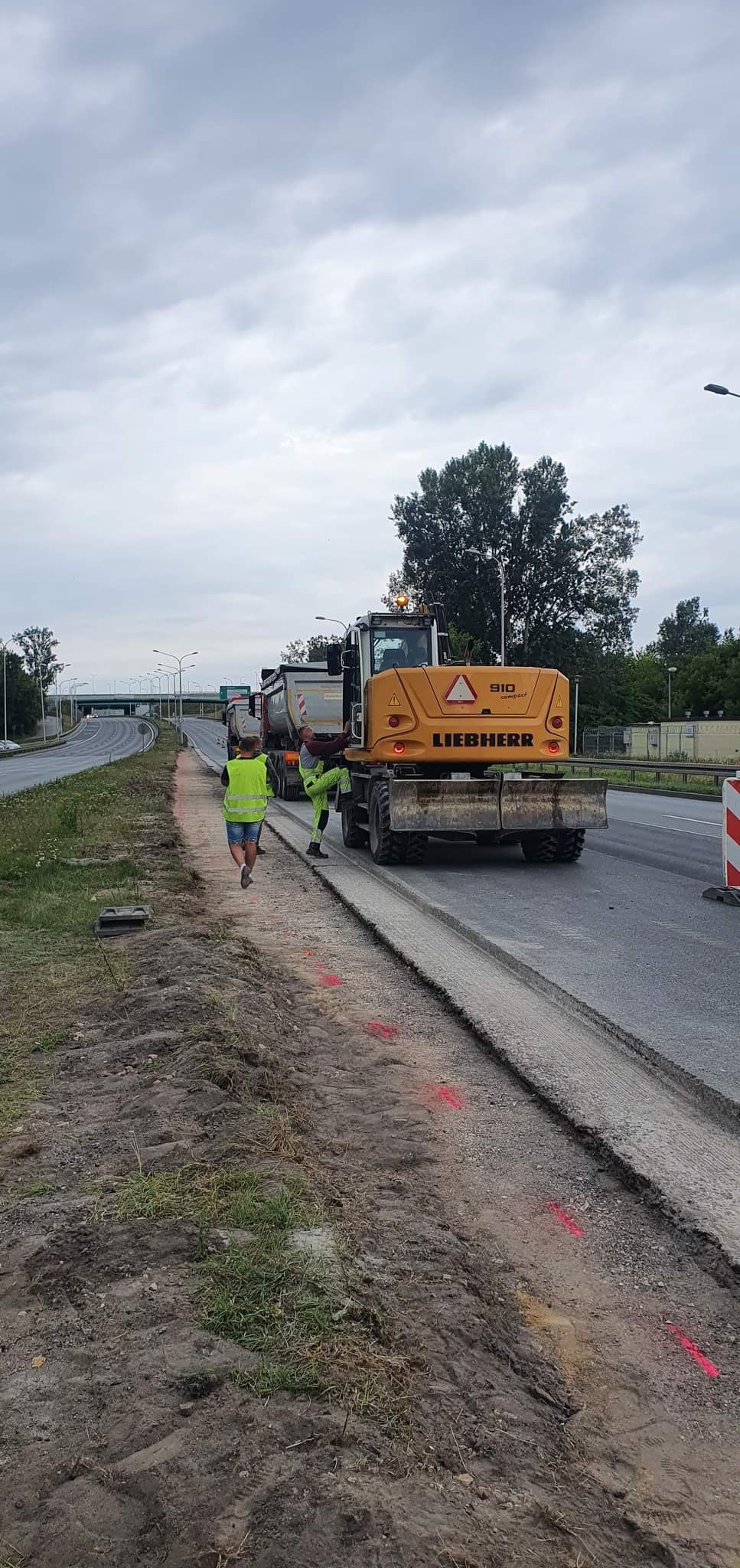 Żółta koparka Liebherr 910 compact przy drodze, pracownicy w odblaskowych ubraniach, wycięty pas asfaltu, obok ciężarówki. Prace drogowe w pochmurny dzień.