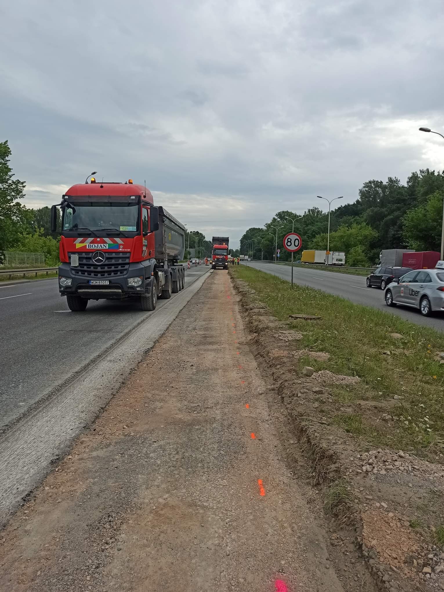 Czerwona ciężarówka na poboczu drogi w trakcie prac ziemnych, widoczne pomarańczowe oznaczenia. W tle ruch samochodów, znak ograniczenia prędkości do 80 km/h.