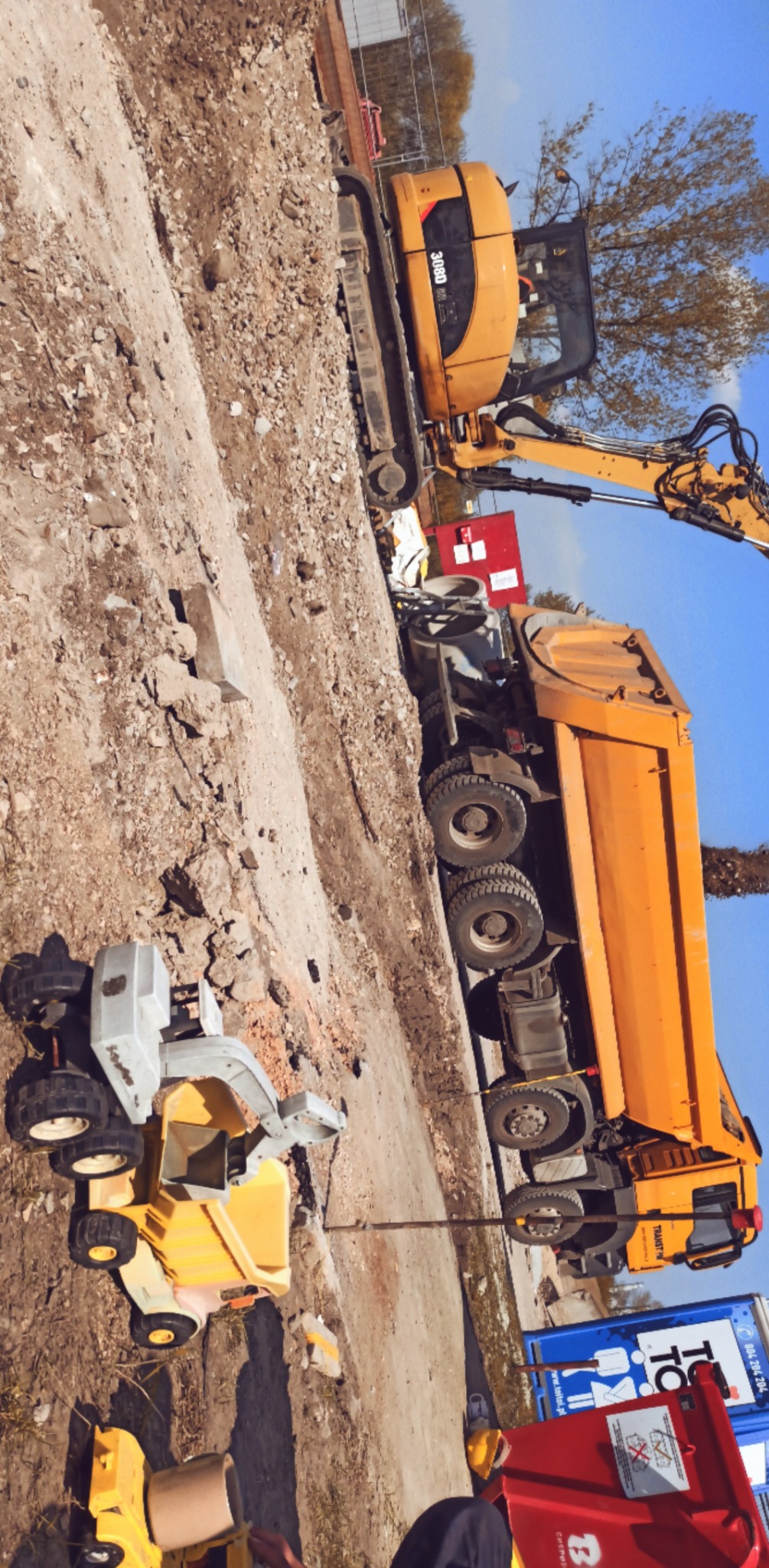 Żółta koparka i wywrotka na placu budowy, z zabawkami budowlanymi w pierwszym planie. W tle widoczne niebo i drzewa. Przekrzywione ujęcie.