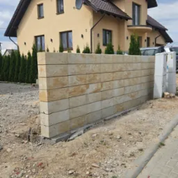 Nowoczesny, niski murek z bloków piaskowca przed domem z beżową elewacją i brązowym dachem. Widoczne elementy instalacji. Świeżo zakończone prace.