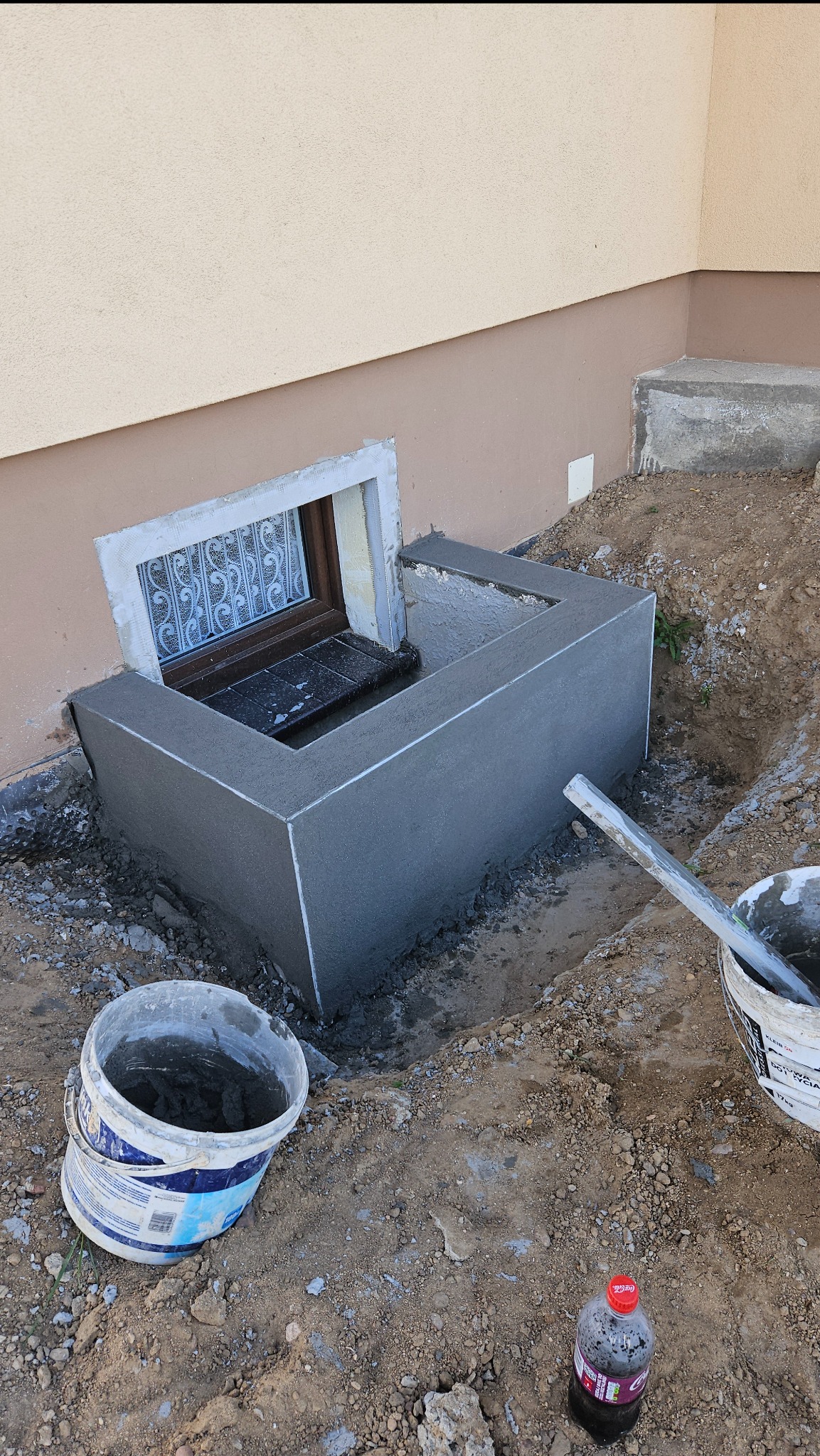 Świeżo wykonana betonowa obudowa okna piwnicznego, z widocznymi śladami zaprawy i narzędziami, na tle ziemi i fragmentu elewacji budynku. Detal wykończenia.
