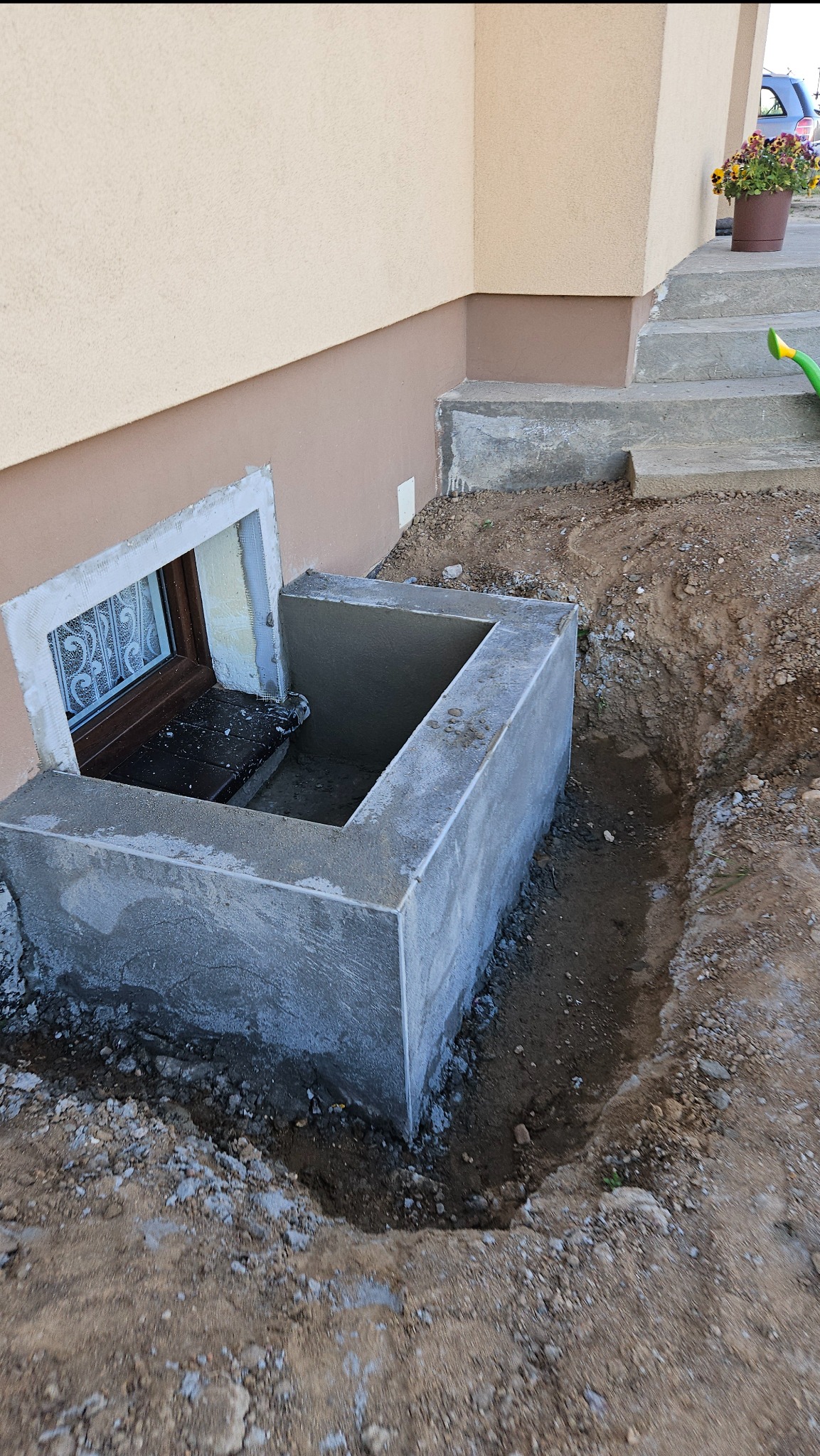 Wykonanie betonowej opaski okiennej przy oknie piwnicznym, widoczne świeże prace murarskie i wykop ziemny. Okno z ozdobną kratą, ściana budynku w dwóch odcieniach beżu.