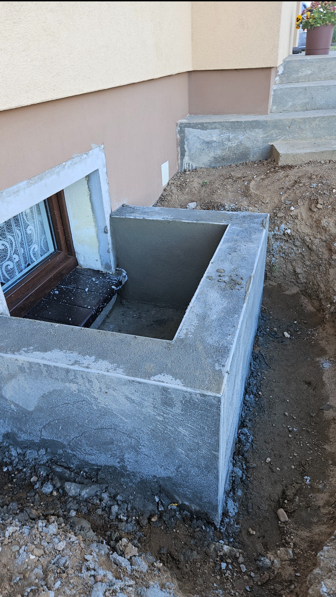 Wykończona betonowa studzienka okienna przy piwnicy, z widoczną wnęką, częściowo obsypana ziemią. Okno z białą ramą i brązowymi szprosami. Schody betonowe w tle.