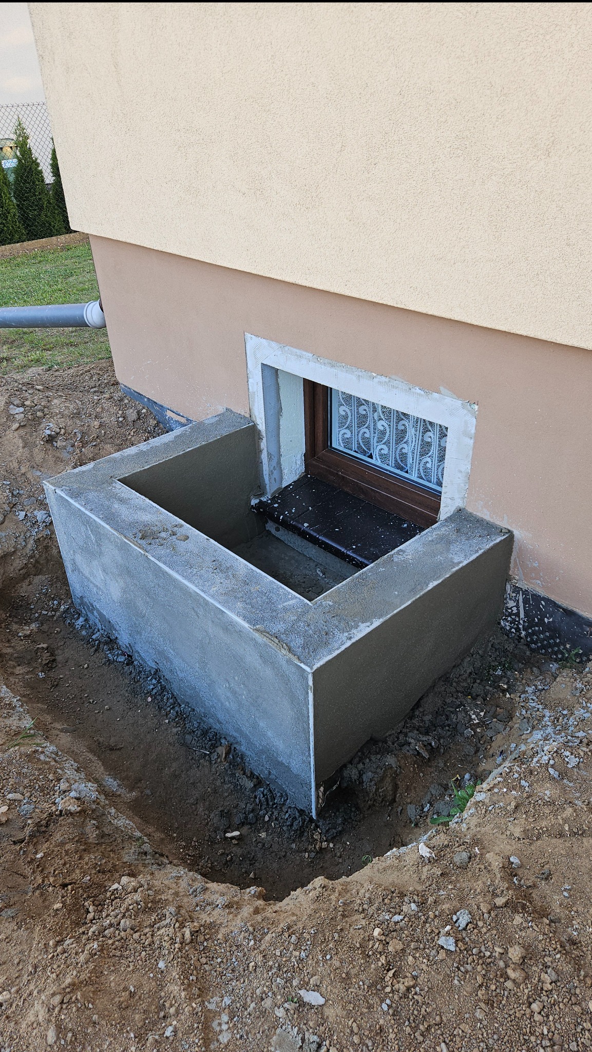 Betonowa studzienka okienna przy elewacji budynku, widok z góry. Okno z kratą w brązowej ramie. Wykopana ziemia wokół studzienki.