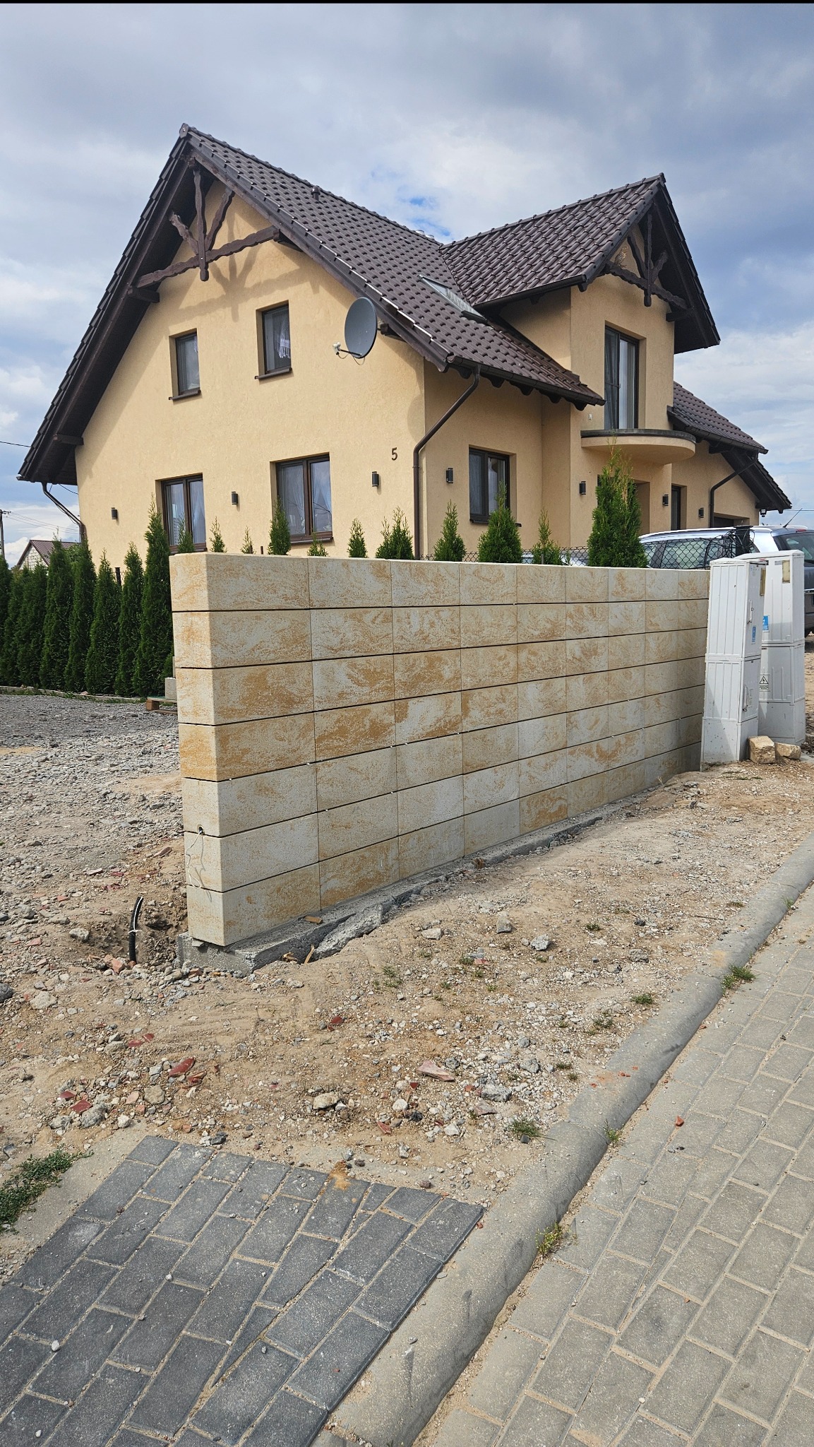 Nowoczesny, niski murek z bloków piaskowca przed domem z beżową elewacją i brązowym dachem. Widoczne elementy instalacji. Świeżo zakończone prace.