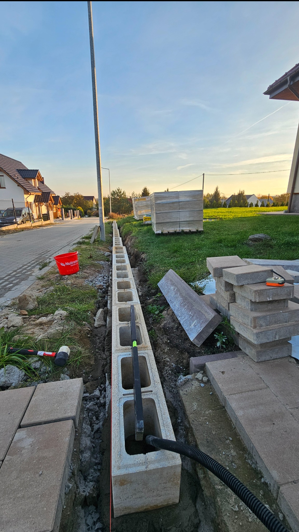 Budowa ogrodzenia z bloczków betonowych z widoczną instalacją wewnątrz, narzędzia pracy i kostka brukowa w tle, w słoneczny dzień.