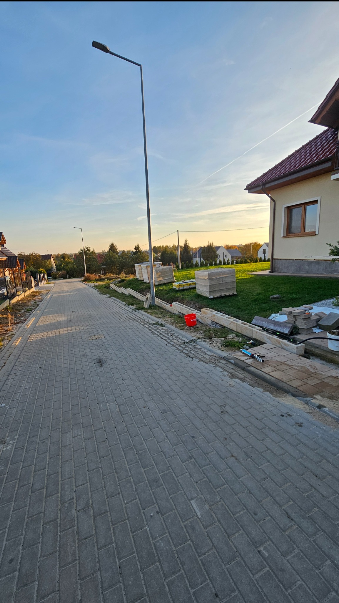 Układanie obrzeży chodnika z kostki, widoczne bloczki betonowe i narzędzia. Praca w toku przy domu jednorodzinnym w słoneczny dzień.