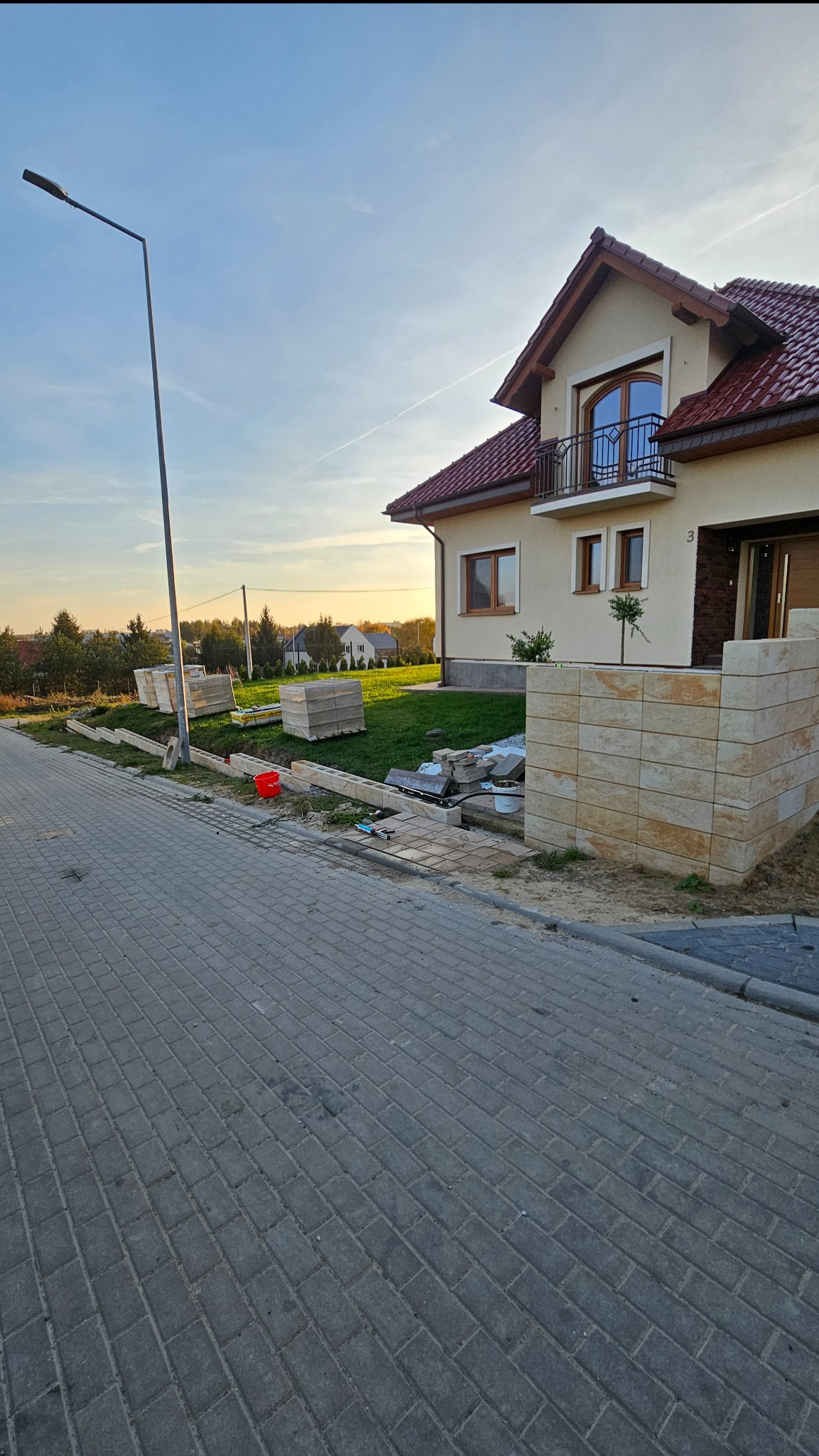 Budowa ogrodzenia z piaskowych bloków przy domu z czerwonym dachem. Na trawie palety z materiałem, widoczne narzędzia i kostka brukowa. Słup oświetleniowy z lewej.