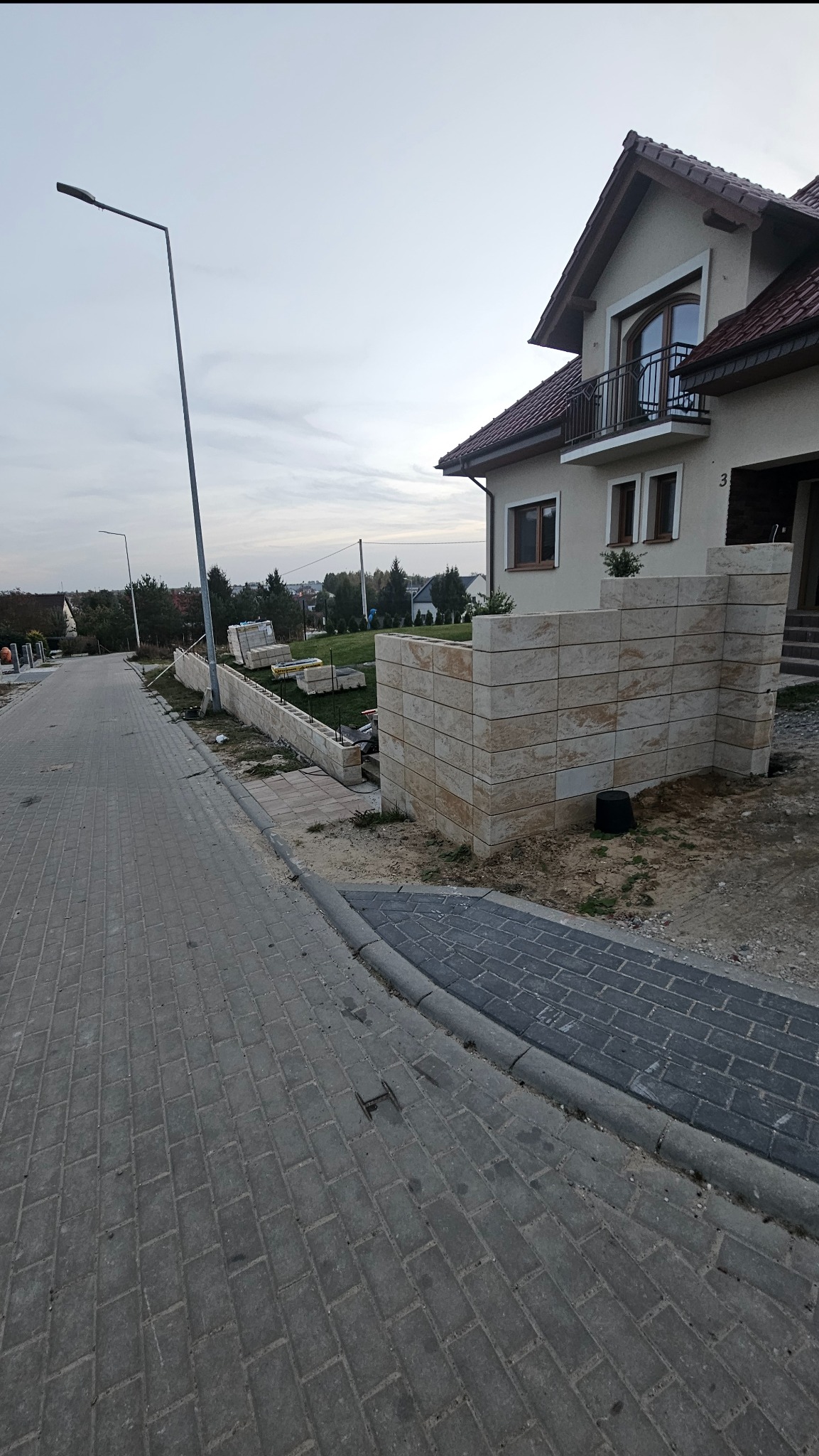 Wznoszenie ogrodzenia z bloków piaskowca przy posesji. Widoczne elementy konstrukcyjne i materiały budowlane. Prace ziemne w toku.