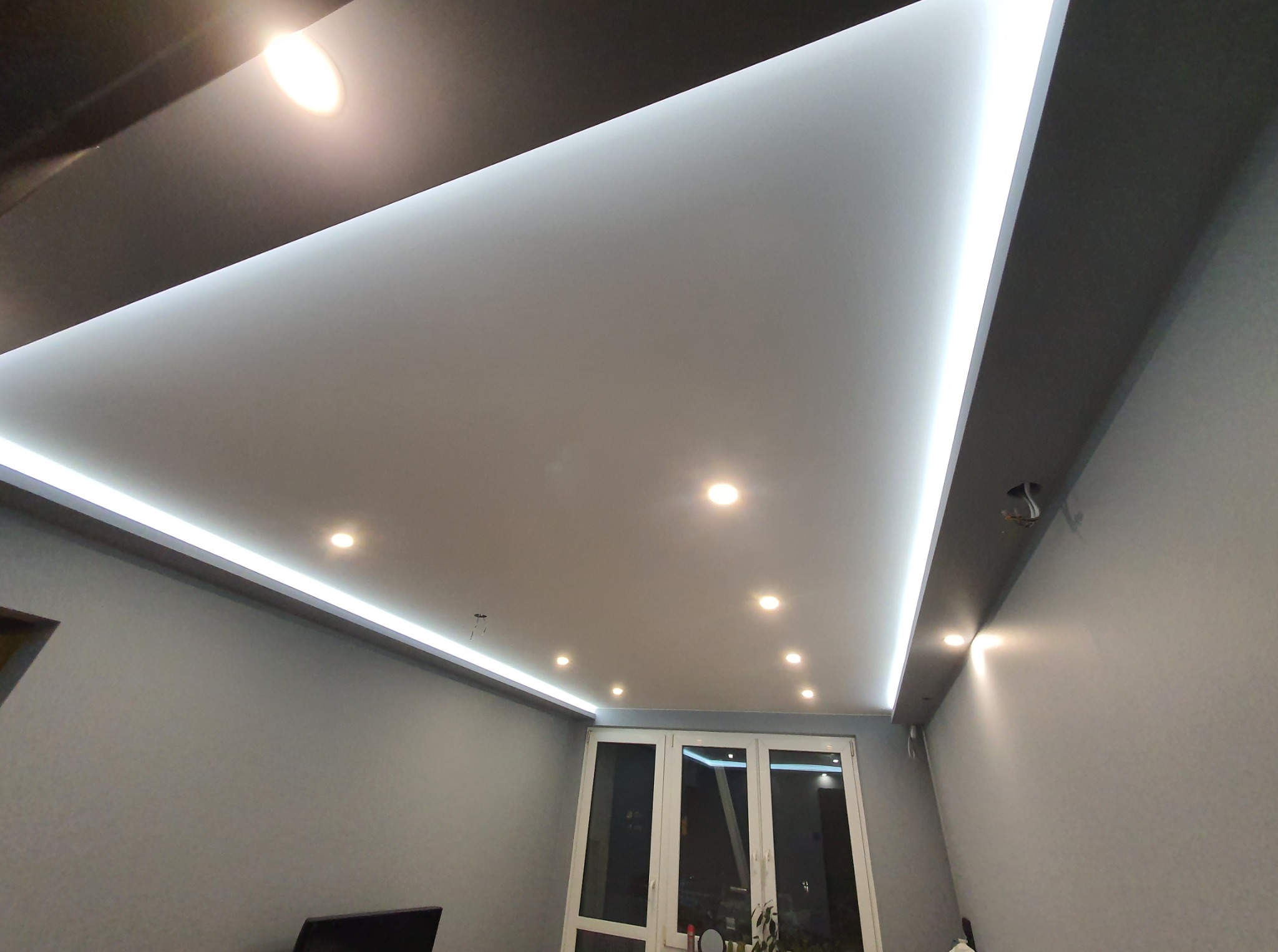 Nowoczesny sufit podwieszany z oświetleniem LED wzdłuż krawędzi i punktowymi lampami. Widok z dołu na szare ściany i okno balkonowe.