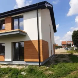 Nowoczesny dom jednorodzinny z białymi ścianami i drewnopodobną elewacją, czarne rynny, trawnik, błękitne niebo z chmurami w tle. Ujęcie z dołu, podkreślające bryłę budynku.