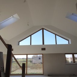 AZ Budowa - Wnętrze poddasza po remoncie: białe ściany i sufit, okna dachowe i panoramiczne z widokiem na zewnątrz. Widoczne przewody elektryczne, drabina i surowa podłoga.