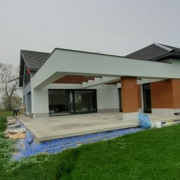 AZ Budowa - Nowoczesny dom jednorodzinny z płaskim dachem i dużymi przeszkleniami, taras w trakcie wykończenia, widoczne elementy izolacji fundamentów.