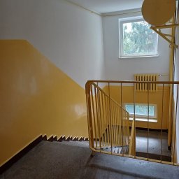 AZ Budowa - Klatka schodowa w bloku pomalowana na żółto. Widoczne schody, ściany, okna i grzejnik. Ujęcie z góry, z naciskiem na kolorystykę i perspektywę.
