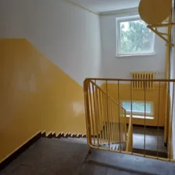 Klatka schodowa w bloku pomalowana na żółto. Widoczne schody, ściany, okna i grzejnik. Ujęcie z góry, z naciskiem na kolorystykę i perspektywę.