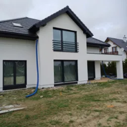 Nowoczesny dom z białymi ścianami i czarnym dachem, widoczne niebieskie węże do izolacji. Okna zabezpieczone folią, teren wokół domu przygotowany do zagospodarowania.